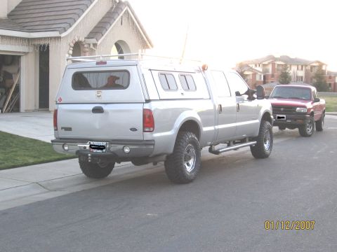 2004 Ford F-250 - A silver pickup truck with a camper shell is parked on the street, with a red pickup truck behind it.