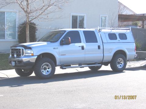 2004 Ford F-250 - This silver 2004 Ford F-250 Super Duty crew cab pickup truck is equipped with a camper shell and roof rack, seen from the side.