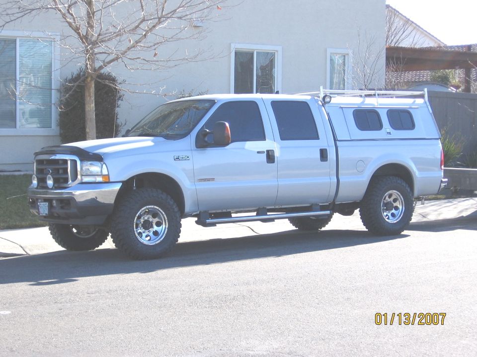 Lifted silver Ford F-250 crew cab with camper shell, front 3/4 view at a curb