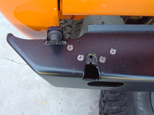Orange vehicle body panel above black metal bumper with visible mounting hardware and tire tread below