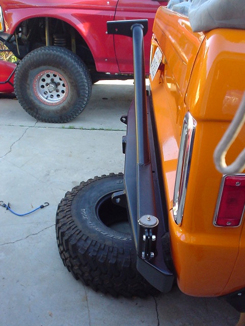 1977 Ford Bronco - Two off-road vehicles are parked side-by-side, with the rear of an orange SUV and the front of a red truck visible.