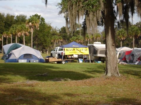 1989 Ford Bronco - A campsite scene tents, an RV, and a pickup truck on a trailer, with palm trees and Spanish moss overhead.