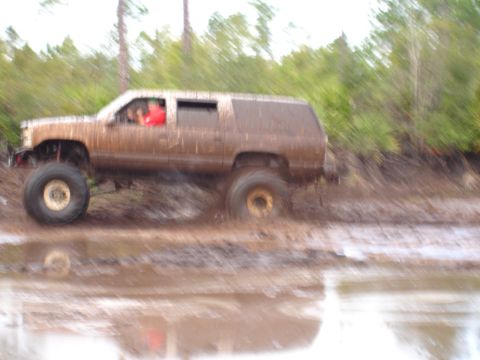 1989 Ford Bronco - A lifted SUV navigates a muddy off-road course, kicking up water and mud.