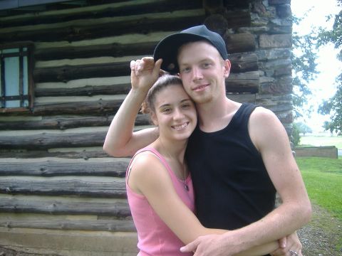 2001 BMW 3 Series - A couple poses for a photo in front of a rustic log cabin with trees in the background.