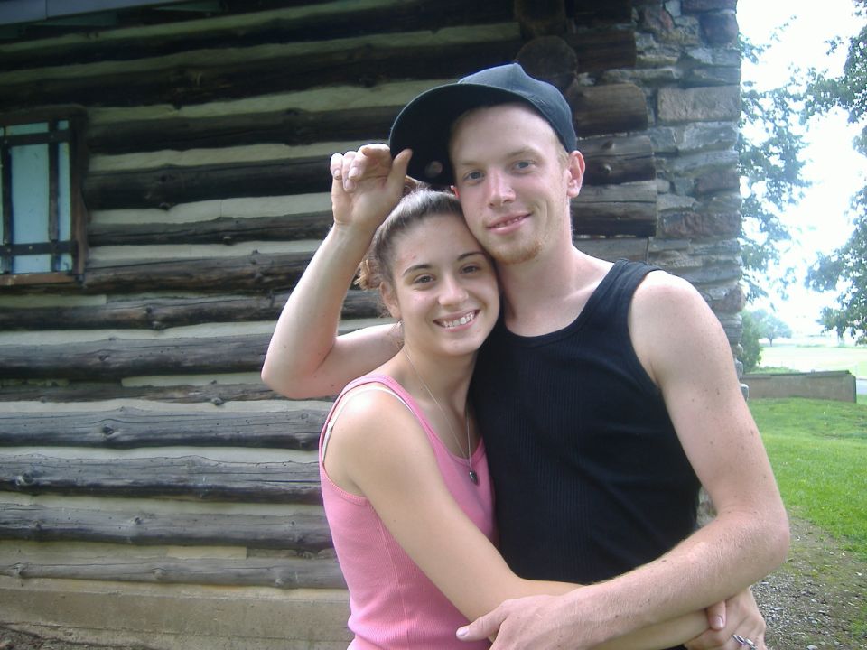 Couple embracing in front of a log cabin, man wearing a black tank top and baseball cap, woman in a pink tank top