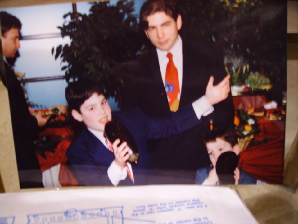 Three people in suits, two boys holding dark objects, at a formal gathering with food on tables