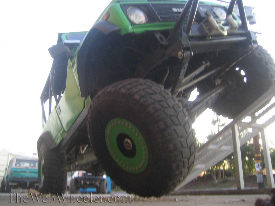 Green off-road vehicle with large tires on a ramp, front view. Another vehicle is visible in the background