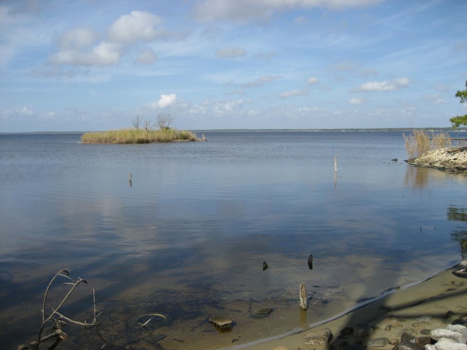 Calm blue water with a small island and reeds under a cloudy sky
