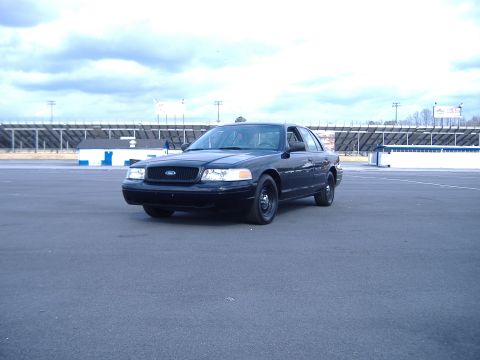 2001 Ford Crown Victoria - This black 2001 Ford Crown Victoria is seen from a three-quarter front view, parked on an asphalt surface with grandstands visible in the d…