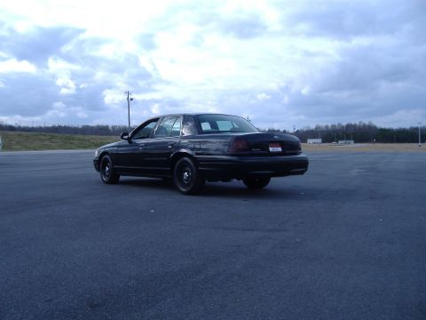2001 Ford Crown Victoria - A black 2001 Ford Crown Victoria is seen from the rear in an open lot under a cloudy sky.