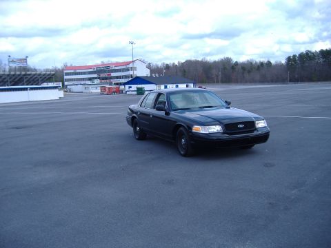 2001 Ford Crown Victoria - A black 2001 Ford Crown Victoria is parked in an empty lot with a racetrack in the background.