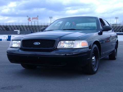 2001 Ford Crown Victoria - A black 2001 Ford Crown Victoria is parked in front of a stadium with a cloudy sky above.