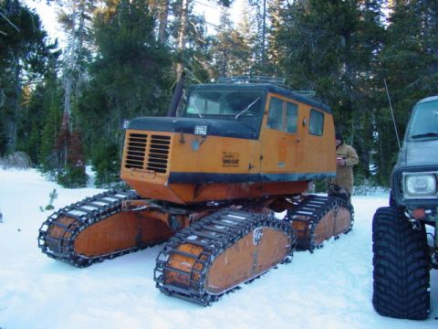 1986 Ford Bronco - A large orange and black tracked vehicle, possibly a snowcat, sits in the snow next to a truck.