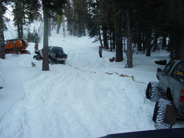 Two off-road vehicles are stuck in deep snow in a wooded area. One vehicle is partially orange and white, the other is a dark SUV. A tow rope connects them