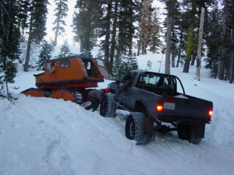 1986 Ford Bronco - A pickup truck and a snow groomer are stuck in the snow in a wooded area.