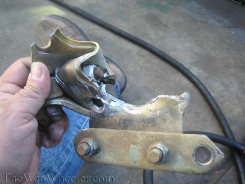 1990 Ford Bronco - hand holds a welded metal bracket above a bolted mounting plate on a garage floor, watermarked TheWebWheeler.com.