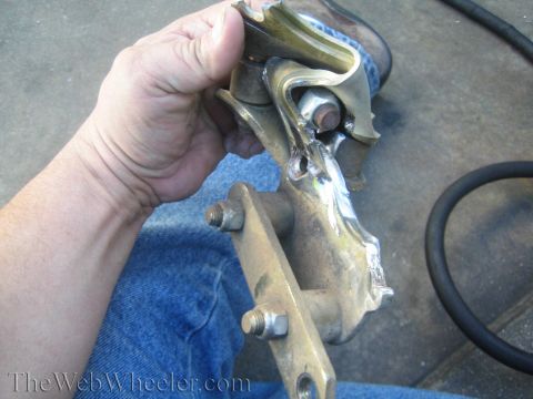 1990 Ford Bronco - hand holds a welded steel bracket assembly over denim-clad legs in a garage, with a hose coiled nearby, watermarked TheWebWheeler.com.