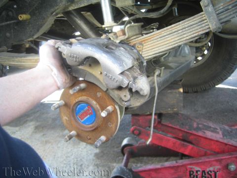 1990 Ford Bronco - hand holds a disc brake caliper beside a wheel hub and leaf spring under a raised truck on a jack, watermarked TheWebWheeler.com.