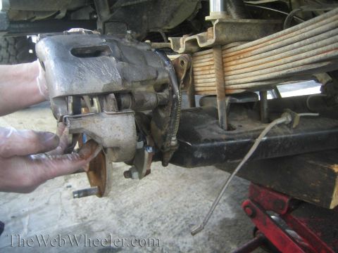 1990 Ford Bronco - hand holds a disc brake caliper next to a leaf spring and frame under a jacked-up vehicle, watermarked TheWebWheeler.com.