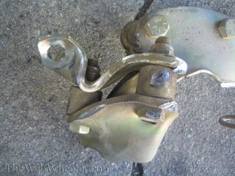 1990 Ford Bronco - Close-up of a yellow zinc-plated steering knuckle or bracket assembly with bolts on a concrete floor, watermarked TheWebWheeler.com.