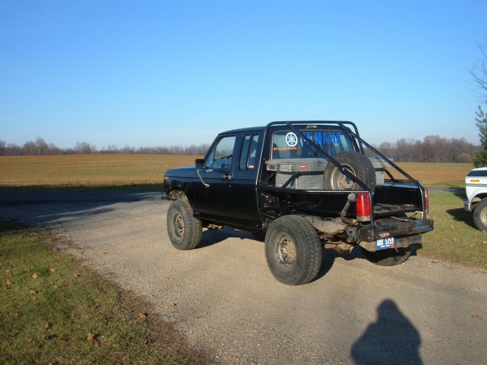 Black lifted extended-cab Ford pickup with cut bedsides, roll bar and toolbox on a gravel road