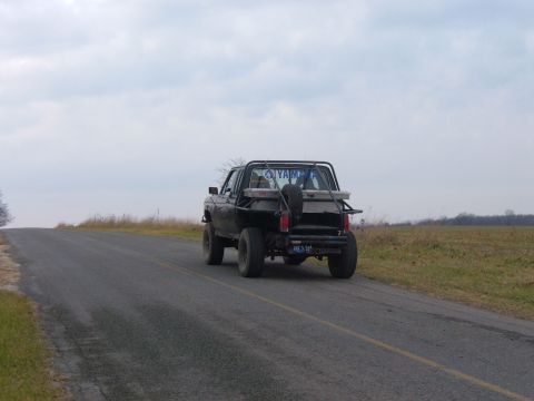1985 Ford F-150 - A modified pickup truck with a spare tire mounted on the back drives down a paved road under a cloudy sky.