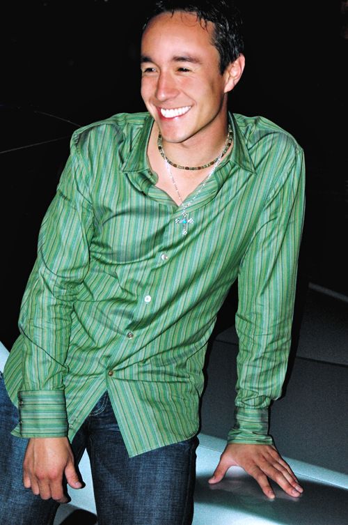Man in green striped shirt and jeans leans on the hood of a silver car