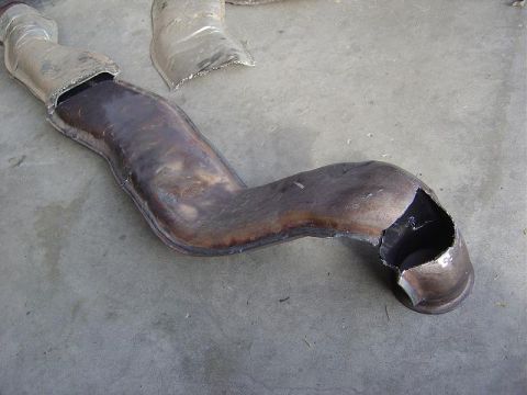 1986 Ford Bronco II - A damaged exhaust pipe lies on a concrete surface, showing a significant hole and signs of wear.