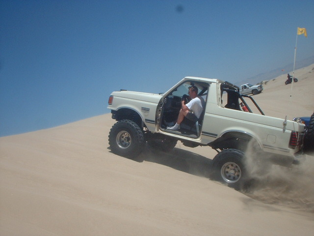 White Ford pickup with doors open driving across sand dunes, driver leaning out