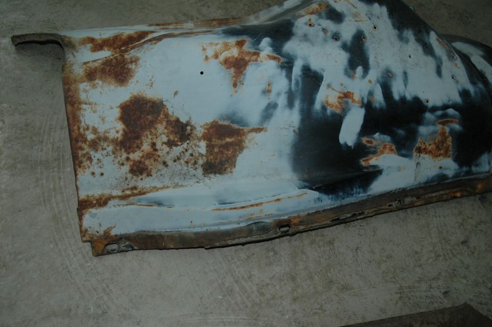 Rusted metal panel with primer and dark undercoating, showing significant corrosion