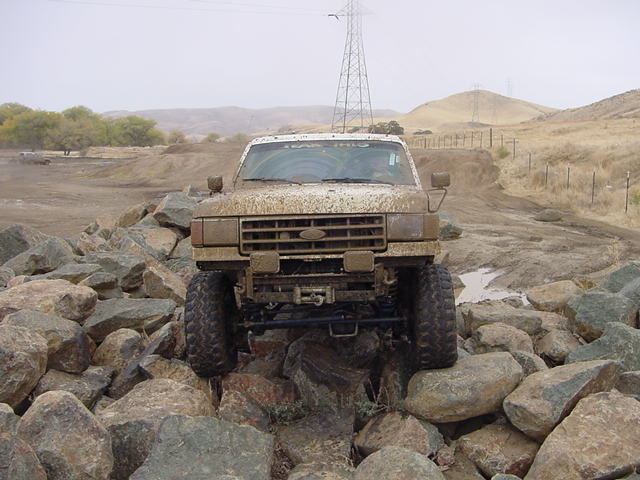 Mud-covered lifted Ford Bronco crawling over rocks, front view with winch and big tires