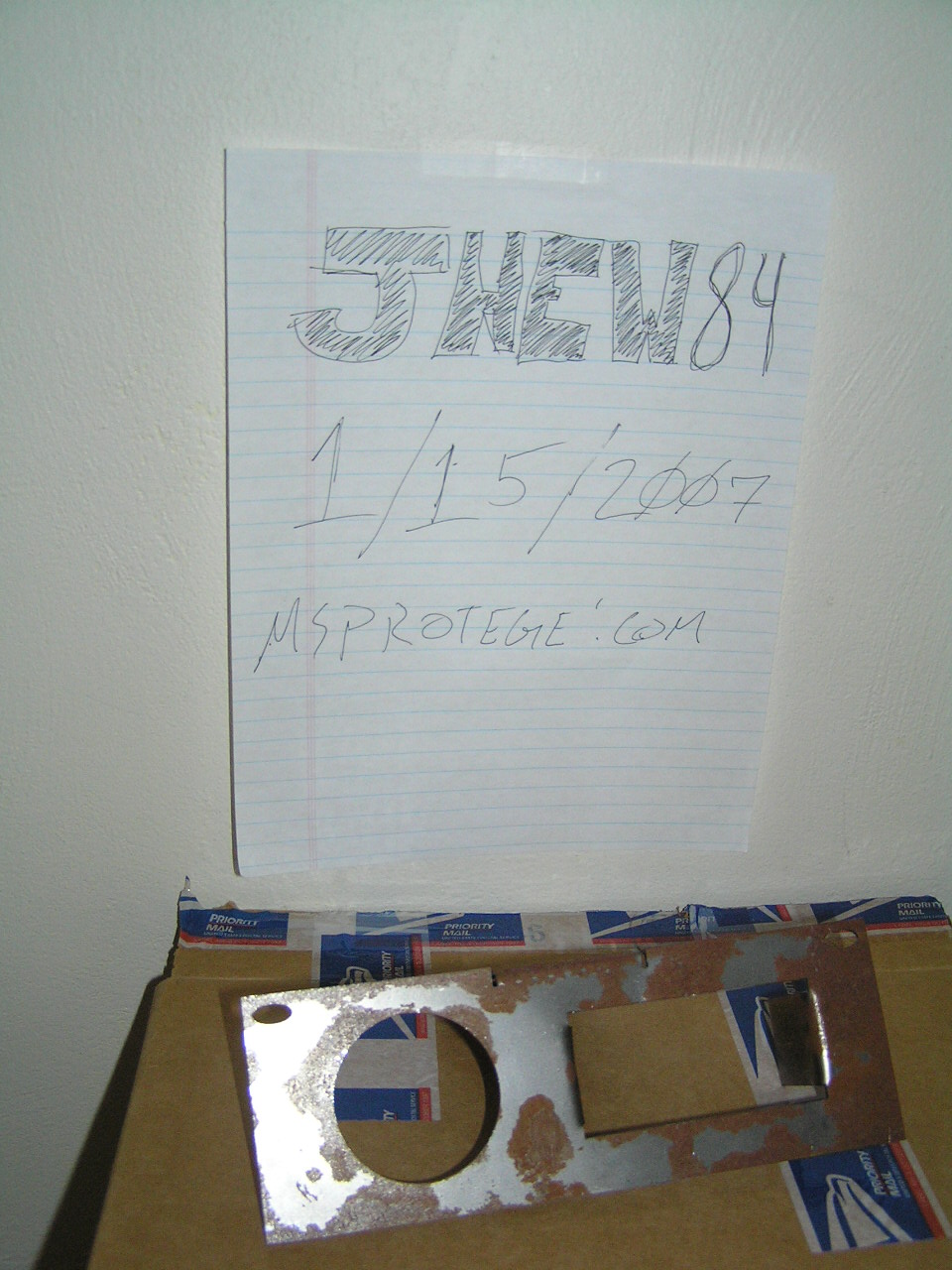 Rusty metal plate with round and rectangular holes on a Priority Mail box, handwritten ID sign above.