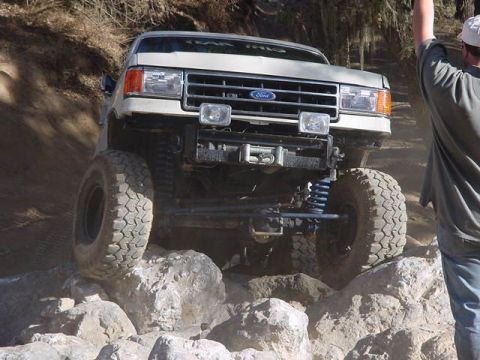 1991 Ford Bronco - A white Ford Bronco is shown off-roading over large rocks, demonstrating its capabilities.