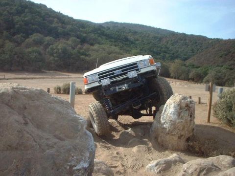 1991 Ford Bronco - A white Ford Bronco is shown off-roading over large rocks on a sunny day.