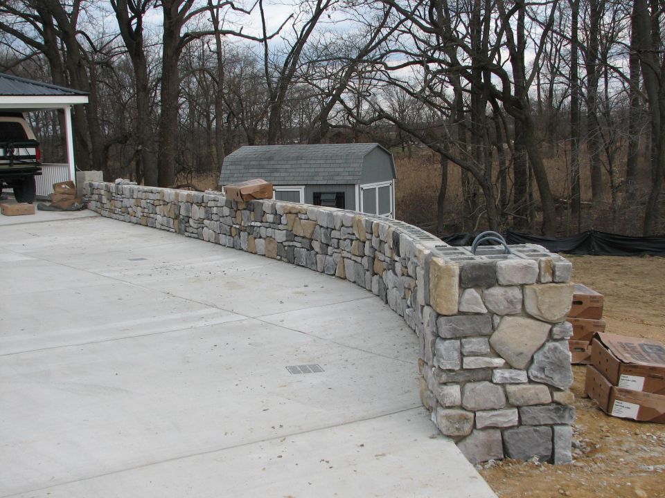 Curved stone veneer retaining wall along a new concrete driveway with storage shed behind