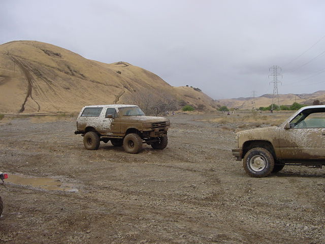 Mud-covered Ford Bronco and Chevy pickup parked on a gravel flat below dry hills