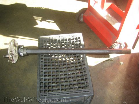 1990 Ford Bronco - removed vehicle axle shaft with its hub flange laid across a black crate beside a red jack stand on a garage floor, watermarked TheWebWheel…