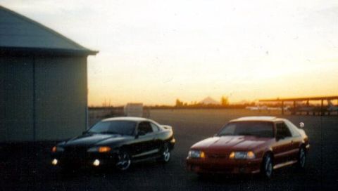 1991 Ford Bronco - Two coupes are parked on an asphalt lot during sunset, with a building to the left and a distant horizon.