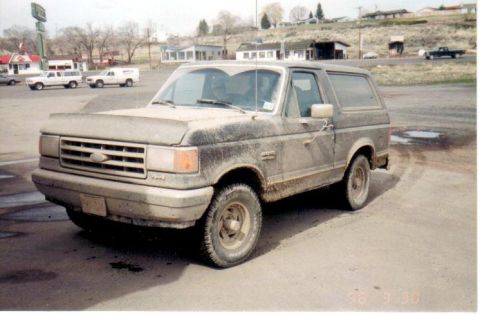 1991 Ford Bronco - A dusty 1991 Ford Bronco sits in a parking lot with buildings and a hill in the background.