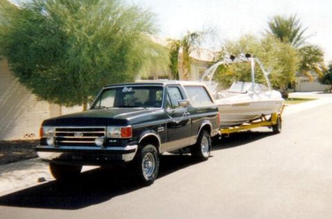 1991 Ford Bronco - A dark green 1991 Ford Bronco is seen towing a boat on a trailer down a suburban street.