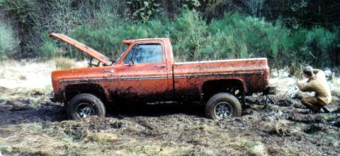1991 Ford Bronco - A person attempts to winch an orange pickup truck out of deep mud, with the hood of the truck open.