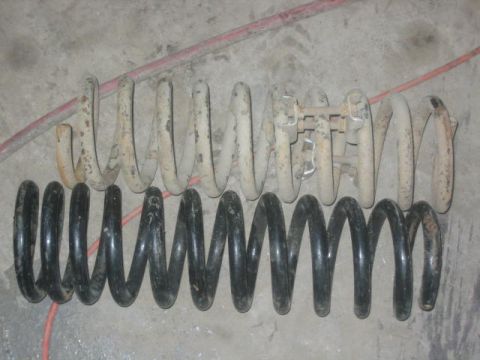 1979 Ford F-150 - Two sets of vehicle suspension springs are laid out on a concrete surface, one set appears new and black, the other is old and rusty.