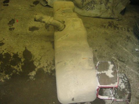 1979 Ford F-150 - An exhaust system part rests on a grimy floor, with two red chairs nearby.