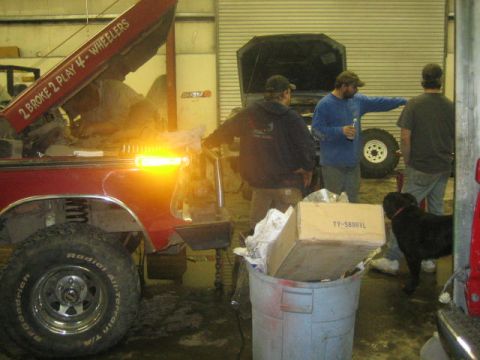 1979 Ford F-150 - A group of people are gathered around a red pickup truck in a garage, with another vehicle in the background.