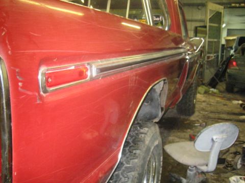 1979 Ford F-150 - Rear quarter panel of a red 1979 Ford F-150 shows chrome trim and signs of wear, possibly in a repair shop.