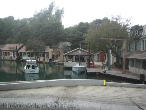 1990 Ford Bronco - movie-set harbor lagoon with small boats and a fake shark suspended from a wooden frame near rustic dock buildings.