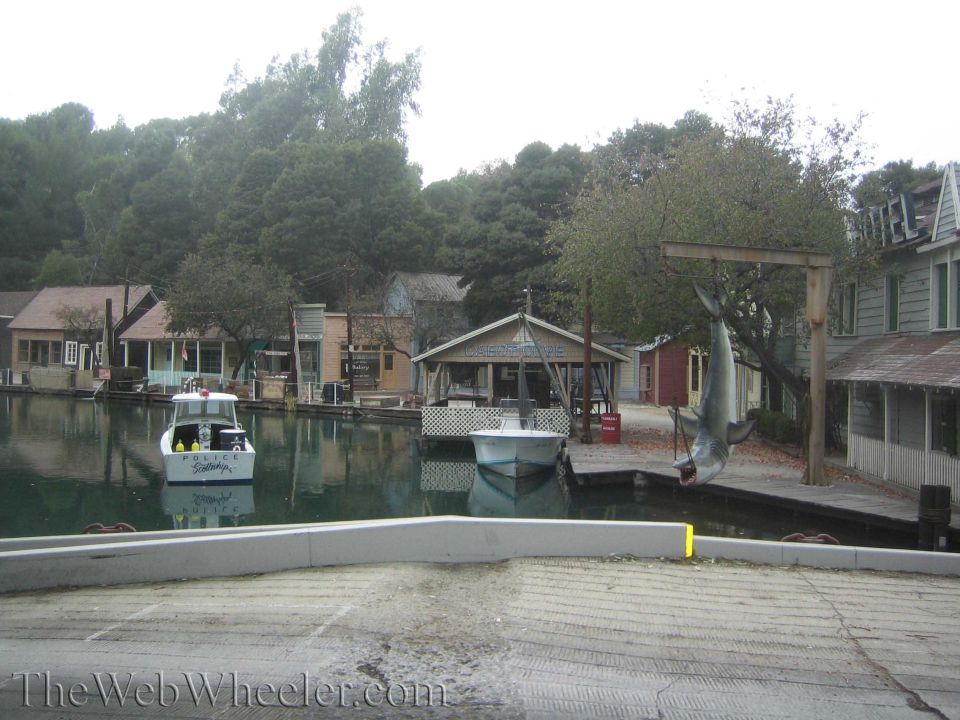 Small boats on a green lagoon with a mechanical shark hung from a wooden gantry beside docks