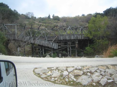 1990 Ford Bronco - dilapidated wooden trestle bridge sags across a dry brushy gully, seen past a vehicle side mirror and a rock-lined curb.