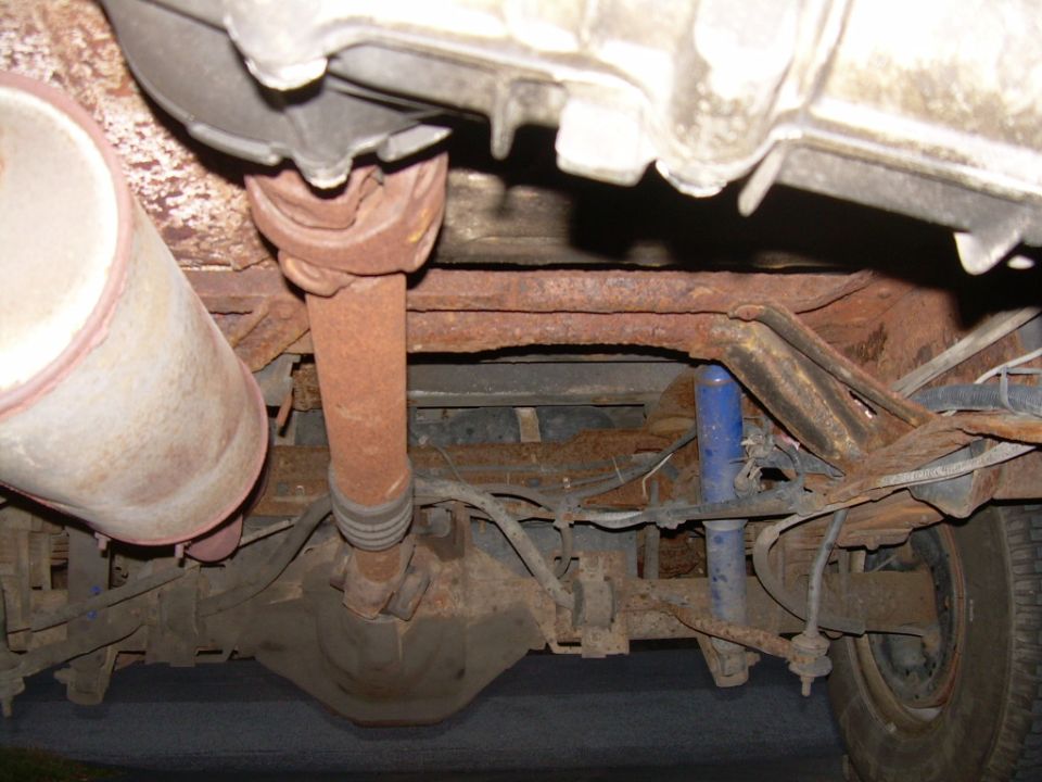 Undercarriage of a rusty vehicle showing a driveshaft, differential, exhaust, and suspension components
