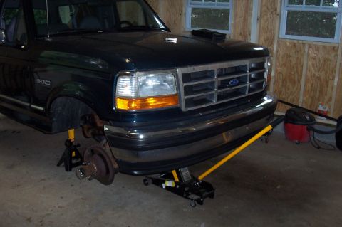 1994 Ford Bronco - Front end of a black Ford Bronco is raised on jack stands in a garage, with the front passenger wheel removed.